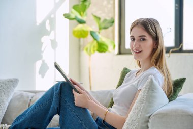 Evdeki kanepede oturup kameraya bakan dijital tabletli, gülümseyen güzel sarışın kadının portresi. Gençlik, rahatlık, rahatlık, rahatlık, yaşam tarzı konsepti