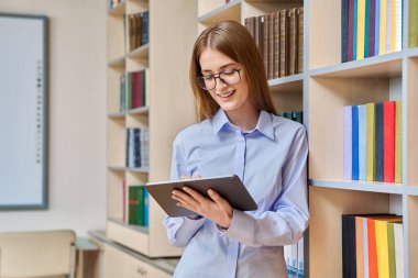 17-18 yaşlarında ciddi bir genç kızın portresi. Sınıfın kütüphanesinde dijital tablet kullanıyor. Gözlüklü sarışın genç kadın, eğitim kitaplarıyla dolu raflar.