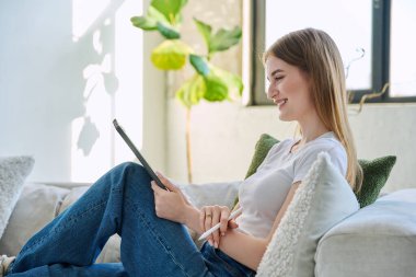 Genç ve güzel bir kız öğrenci dijital tabletle online sohbet konferansı yapıyor. Evde, kanepede oturuyor. İletişim öğrenme teknolojileri gençlerin video bloglarında boş zamanlarında çalışıyor