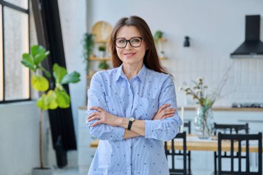 Kendinden emin, orta yaşlı, gülümseyen, güzel bir kadın ev içinde kollarını kavuşturarak kameraya bakıyor. Başarı, güven, yaşam tarzı, olgun insanlar konsepti