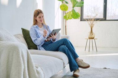 Evdeki kanepede dijital tablet kullanan genç bir kadın. Cihazlı kadın üniversite öğrencisi, rahatlama eğitimi, eğlence iletişimi eğitimi teknolojisi, yaşam tarzı gençlik konsepti