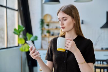 Elinde bir bardak içkiyle evde duran akıllı telefonu kullanan genç bir kadın. Mesajlaşan genç kız gezici uygulamaları okuyor. İnternet teknolojisi. Boş zaman iletişim yaşam tarzı eğitimi için internet hizmetleri.