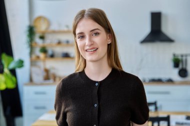 Güzel, gülümseyen, sarı saçlı bir kızın portresi. Kameraya bakıyor, evin arka planında. Başarılı çekici genç kız öğrenci üniversite yaşı, yaşam tarzı, gençlik konsepti