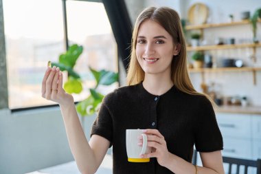 Genç, güzel, gülümseyen bir kadın balık yağı Omega 3 ile sarı kapsül hapı alıyor, evde duruyor. Sağlıklı beslenme, diyet, vitaminler, gıda takviyeleri, sağlık hizmetleri kavramı