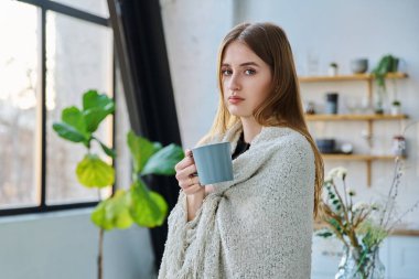 Omuzlarında sıcak battaniyesi olan genç ve sakin bir kadın pencerenin yanındaki kameraya bakıyor. Sonbahar kış mevsimi, soğuk hava, ev konforu rahatlama konsepti