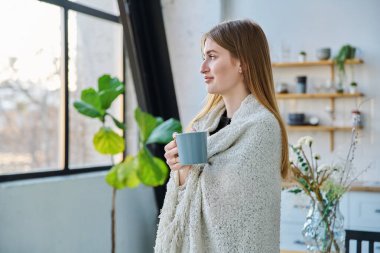 Güzel, gülümseyen, mutlu, sakin bir kadın omuzlarında sıcak bir battaniyeyle pencereden dışarı bakan bir kupa tutuyor. Sonbahar kış mevsimi, soğuk hava, ev konforu rahatlama konsepti