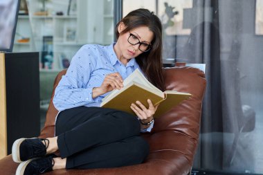 Orta yaşlı, ciddi, kitap okuyan bir kadın evdeki koltukta oturuyor. Boş zaman, hobi, okuma, kurgu, iş, eğitim, edebiyat.