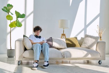 20 yaşında genç bir adam akıllı telefon kullanarak kanepede oturuyor. Kıvırcık gözlüklü erkek evde cep telefonuyla dinleniyor, yaşam tarzı teknoloji gençliği kavramı.