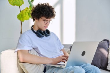 Evdeki kanepede dizüstü bilgisayar kullanan yakışıklı bir erkek portresi. 20 yaşında, bilgisayar başında daktilo yazan gözlüklü, kıvırcık bir öğrenci. Yaşam tarzı, eğitim, teknoloji, gençlik kavramı