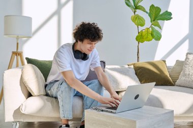 Evdeki kanepede dizüstü bilgisayar kullanan yakışıklı bir erkek portresi. 20 yaşında, bilgisayar başında daktilo yazan gözlüklü, kıvırcık bir öğrenci. Yaşam tarzı, eğitim, teknoloji, gençlik kavramı