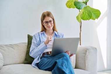 Gözlüklü genç bir bayan bilgisayar kullanarak evdeki kanepede oturup görüntülü sohbet konferansına katılıyor. İletişim öğrenimi için dijital teknolojiler boş zamanlarında çalışır