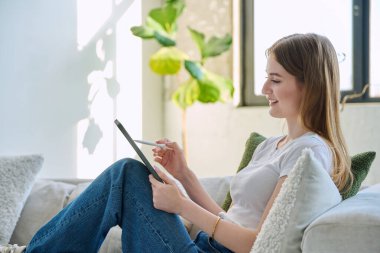Evdeki kanepede dijital tablet kullanan genç bir kadın. Cihazlı kadın üniversite öğrencisi, rahatlama eğitimi, eğlence iletişimi eğitimi teknolojisi, yaşam tarzı gençlik konsepti