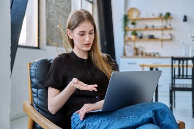 Genç ve güzel bir kadın internette sohbet konferansı için dizüstü bilgisayar kullanıyor. Evde, sandalyesinde oturuyor. İletişim öğrenmek için dijital teknolojiler boş zamanlarında sosyal medya bloglarında çalışır