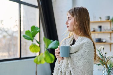 Omuzlarında sıcak battaniyeyle pencereden dışarı bakan güzel, üzgün, ciddi, sakin bir kadın. Sonbahar kış mevsimi, soğuk hava, ev konforu rahatlama konsepti