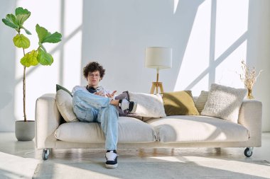 20 yaşında genç bir adam akıllı telefon kullanarak kanepede oturuyor. Kıvırcık gözlüklü erkek evde cep telefonuyla dinleniyor, yaşam tarzı teknoloji gençliği kavramı.