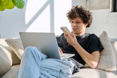 Evdeki kanepede oturan akıllı telefon kullanan, dizüstü bilgisayarı olan yakışıklı, serbest çalışan bir genç. Kıvırcık saçlı ciddi bir adam, modern dijital aletlerle iletişim işi eğitimi alıyor.