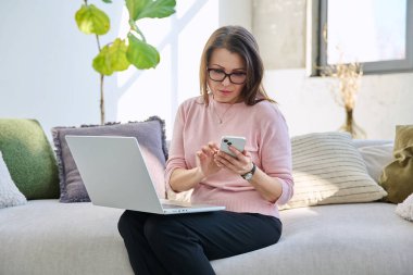 Orta yaşlı bir kadın oturma odasının içinde dizüstü bilgisayarlı akıllı telefonuyla kanepede oturuyor. Olgun kadın iletişim işi için modern dijital cihazlar kullanıyor.