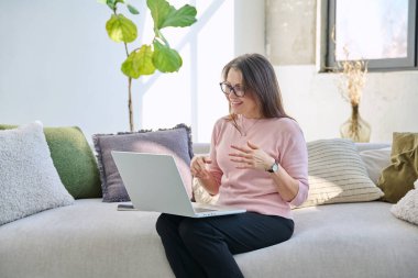 Orta yaşlı bir kadın uzaktan çalışıyor, görüntülü konuşma yapıyor, koltukta dizüstü bilgisayar kullanıyor. Psikoloji oturumu çevrimiçi öğretim koçluğu vlog işi iletişimi