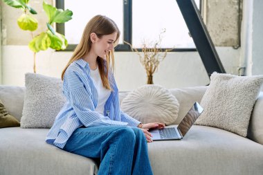 Genç ve güzel bir kadın internette sohbet etmek için dizüstü bilgisayar kullanıyor. Evde, kanepede oturuyor. İletişim öğrenmek için dijital teknolojiler boş zamanlarında sosyal medya bloglarında çalışır