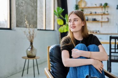 Genç ve güzel bir kadının portresi, gülümseyen üniversite öğrencisi evinde, sandalyesinde oturan pencereye bakıyor. Çekici kadın sarışın, yaşam tarzı, gençlik kavramı.