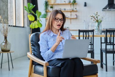 Orta yaşlı bir kadın uzaktan çalışıyor, video konferans görüşmeleri yapıyor, ofis koltuğunda dizüstü bilgisayar kullanıyor. Psikoloji oturumu çevrimiçi öğretim koçluğu vlog işi iletişimi