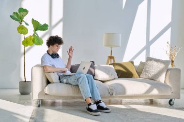 Genç yakışıklı kıvırcık adam internette sohbet etmek için bilgisayarını kullanıyor. Evde, koltukta oturuyor. İletişim öğrenimi için dijital teknolojiler boş zamanlarında sosyal medya bloglarında çalışır