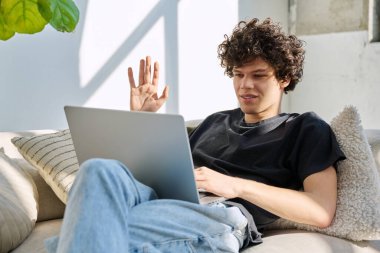 Genç yakışıklı kıvırcık adam internette sohbet etmek için bilgisayarını kullanıyor. Evde, koltukta oturuyor. İletişim öğrenimi için dijital teknolojiler boş zamanlarında sosyal medya bloglarında çalışır