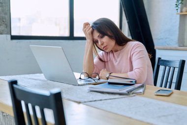 Depresyonda olan, stresli, zihinsel ve fiziksel acı çeken olgun bir kadın bilgisayar laptopuyla masada oturuyor. Sorunlar yalnızlık, baş ağrısı hormon stresi, orta yaşlı kadınların sağlığı