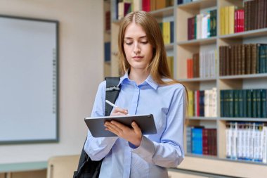 17-18 yaşlarında ciddi bir genç kızın portresi. Sınıfın kütüphanesinde dijital tablet kullanıyor. Gözlüklü sarışın genç kadın, eğitim kitaplarıyla dolu raflar.