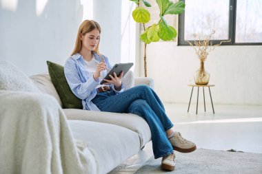 Evdeki kanepede dijital tablet kullanan genç bir kadın. Cihazlı kadın üniversite öğrencisi, rahatlama eğitimi, eğlence iletişimi eğitimi teknolojisi, yaşam tarzı gençlik konsepti