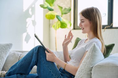 Genç ve güzel bir kız öğrenci dijital tabletle online sohbet konferansı yapıyor. Evde, kanepede oturuyor. İletişim öğrenme teknolojileri gençlerin video bloglarında boş zamanlarında çalışıyor