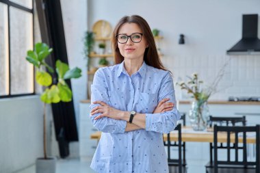 Kendinden emin, orta yaşlı, güzel bir kadın ev içinde kollarını kavuşturmuş kameraya bakıyor. Başarı, güven, yaşam tarzı, olgun insanlar konsepti