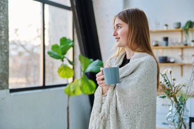 Güzel, gülümseyen, mutlu, sakin bir kadın omuzlarında sıcak bir battaniyeyle pencereden dışarı bakan bir kupa tutuyor. Sonbahar kış mevsimi, soğuk hava, ev konforu rahatlama konsepti