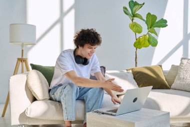 Genç yakışıklı kıvırcık adam internette sohbet etmek için bilgisayarını kullanıyor. Evde, koltukta oturuyor. İletişim öğrenimi için dijital teknolojiler boş zamanlarında sosyal medya bloglarında çalışır