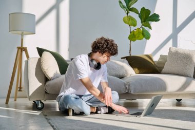 Evdeki kanepenin yanında dizüstü bilgisayarda yazan yakışıklı bir erkek portresi. Kıvırcık, güler yüzlü, 20 yaşında bir öğrenci. Yaşam tarzı, eğitim, teknoloji, gençlik kavramı