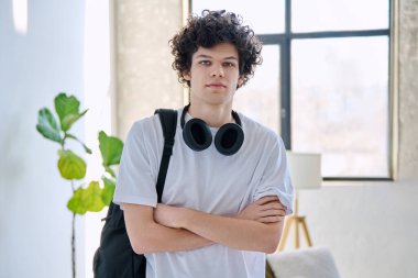 Çapraz kollu, kulaklıklı, sırt çantalı, kendinden emin genç bir adam. Moda tarzında kıvırcık saçlı, kameraya bakan poz veren adam. Gençlik, yaşam tarzı, yirmili yaşlar konsepti