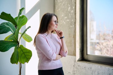 Orta yaşlı sakin, kendinden emin, pencereden dışarı bakan, gülümseyen olgun bir kadın hayal eden, profil manzaralı, kopyalayan bir kadın. Yaşam tarzı, orta yaşlı insanlar konsepti
