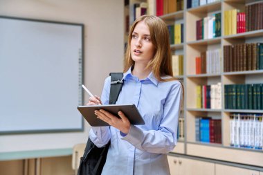 17-18 yaşlarında ciddi bir genç kızın portresi. Sınıfın kütüphanesinde dijital tablet kullanıyor. Gözlüklü sarışın genç kadın, eğitim kitaplarıyla dolu raflar.