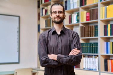 Kollarını kavuşturup kütüphane ofisinde poz veren kendine güvenen yakışıklı genç bir adamın portresi. Başarılı gülümseyen 30 'lu yaşlardaki erkek öğretmen, akıl hocası, eğitim işçisi kameraya bakıyor.