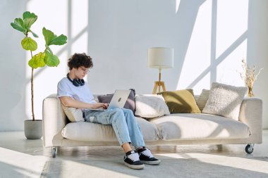 Evdeki kanepede dizüstü bilgisayar kullanan yakışıklı bir erkek portresi. 20 yaşında, bilgisayar başında daktilo yazan gözlüklü, kıvırcık bir öğrenci. Yaşam tarzı, eğitim, teknoloji, gençlik kavramı