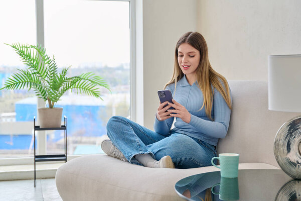 Молодая красивая улыбчивая женщина, сидящая дома в кресле с помощью смартфона, читает, читает. Технологии мобильных приложений досуга коммуникация шопинг исследования блог блог молодежь концепция