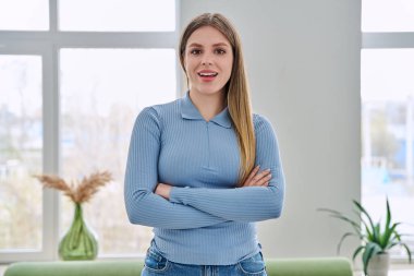 İç mekanda kollarını kavuşturarak poz veren kendinden emin genç bir kadının portresi. Gülümseyen mutlu 20 'li yaşlarda, sarışın, uzun saçlı, kameraya bakan bir kadın. Güzellik sağlık yaşam tarzı gençlik kavramı