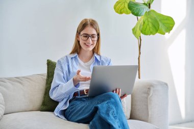 Gözlüklü genç bir bayan bilgisayar kullanarak evdeki kanepede oturup görüntülü sohbet konferansına katılıyor. İletişim öğrenimi için dijital teknolojiler boş zamanlarında çalışır