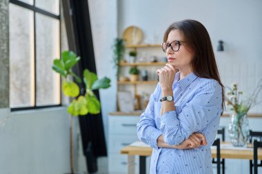Ciddi bir orta yaşlı kadın portresi. Evin içinde pencereden dışarı bakıyor. Duygular, yaşam tarzı, olgun insanlar konsepti