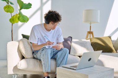 20 yaşında, kulaklıklı genç bir öğrenci. Koltukta dizüstü bilgisayarla oturuyor. Evde ders çalışırken not alıyor. Eğitim, bilgi, teknoloji, gençlik, yaş üniversitesi konsepti