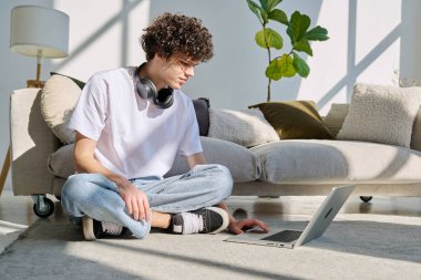 Evdeki kanepenin yanında dizüstü bilgisayarda yazan yakışıklı bir erkek portresi. Kıvırcık, güler yüzlü, 20 yaşında bir öğrenci. Yaşam tarzı, eğitim, teknoloji, gençlik kavramı