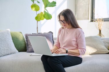 Evdeki kanepede oturup dizüstü bilgisayarda yazı yazan orta yaşlı güzel bir iş kadınının portresi, gevşemiş ciddi bir kadın. Teknoloji, yaşam tarzı, iş eğlencesi, insan konsepti