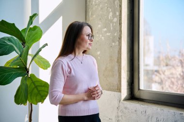 Orta yaşlı sakin, kendinden emin, pencereden dışarı bakan, gülümseyen olgun bir kadın hayal eden, profil manzaralı, kopyalayan bir kadın. Yaşam tarzı, orta yaşlı insanlar konsepti