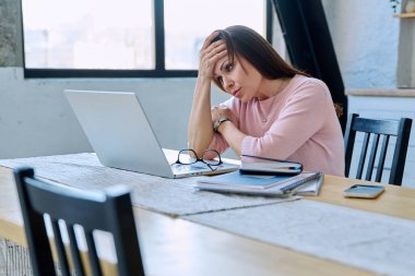 Depresyonda olan, stresli, zihinsel ve fiziksel acı çeken olgun bir kadın bilgisayar laptopuyla masada oturuyor. Sorunlar yalnızlık, baş ağrısı hormon stresi, orta yaşlı kadınların sağlığı