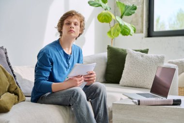 Ciddi bir ergen erkek öğrenci evinde oturmuş, dizüstü bilgisayarla kameraya bakıyor. Eğitim, teknoloji, ergenlik kavramı